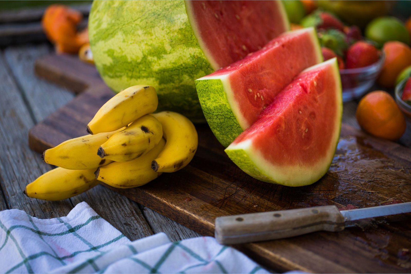Kalio turtingi bananai guli ant pjaustymo lentelės šalia L-citrulino turtingo pjaustyto arbūzo.